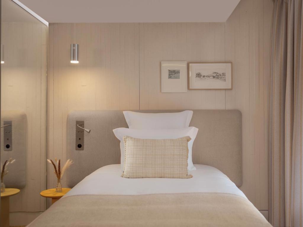 Urban seminar hotel single bedroom with a single bed dressed in white linen and a beige woven cushion against a light wood-panelled upholstered headboard wall, a chrome reading sconce and two small landscape sketch frames above, a small round yellow bedside table with dried grasses on the left, and a glazed bathroom partition visible on the left.