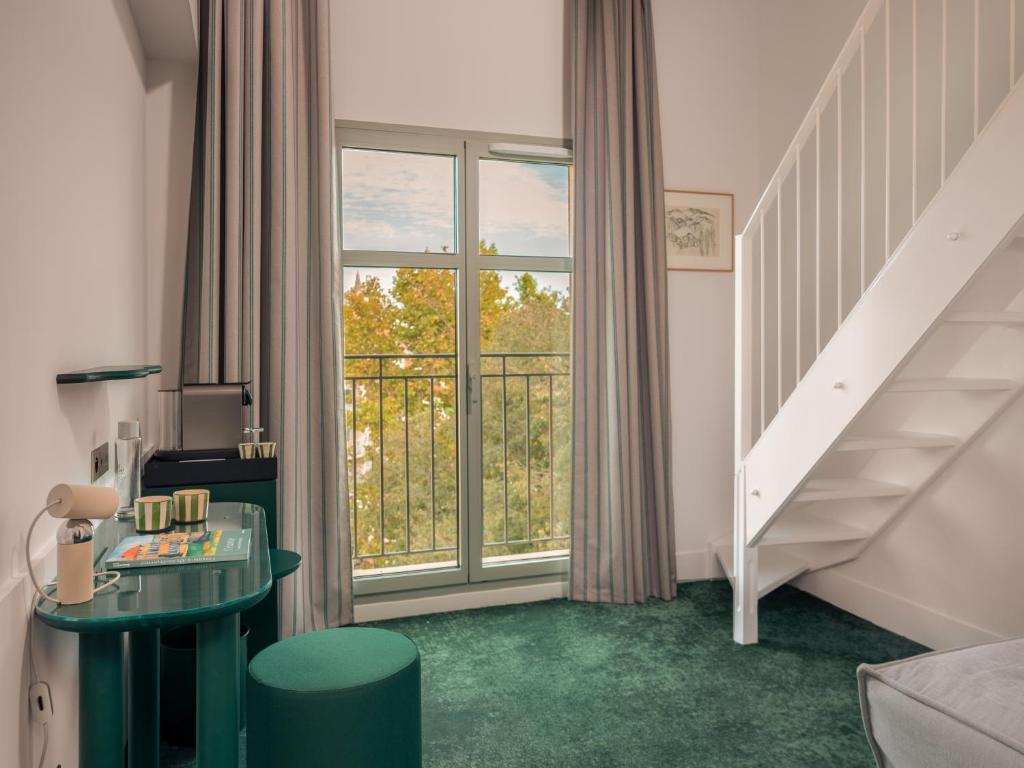 Hotel group accommodation duplex room with a deep green carpet, a green round side table with striped mugs, a small lamp and a magazine on the left, a green velvet pouf in the foreground, a large window with grey striped curtains overlooking green trees and a balcony with a metal railing, a white wooden staircase leading to the upper level on the right, and a framed artwork on the white wall.