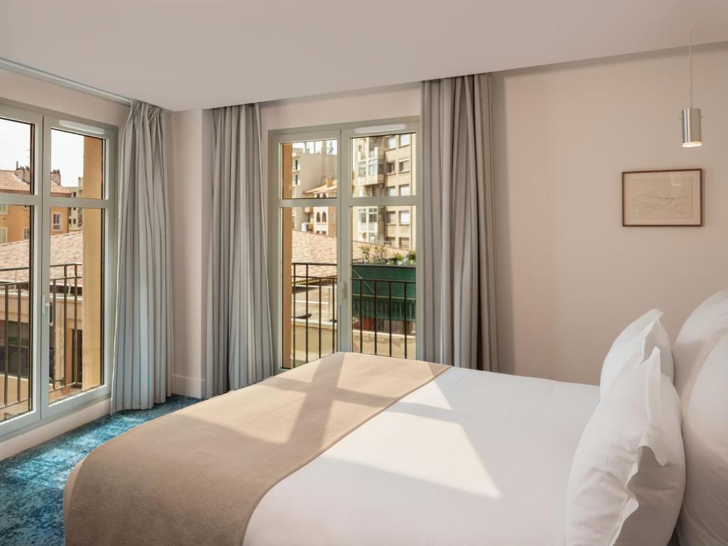 Urban seminar hotel bedroom with a double bed dressed in white linen and a beige runner on a blue patterned carpet, grey striped curtains framing two large windows with a balcony overlooking Aix-en-Provence rooftops and buildings, a chrome pendant light and a small framed artwork on the beige wall to the right.