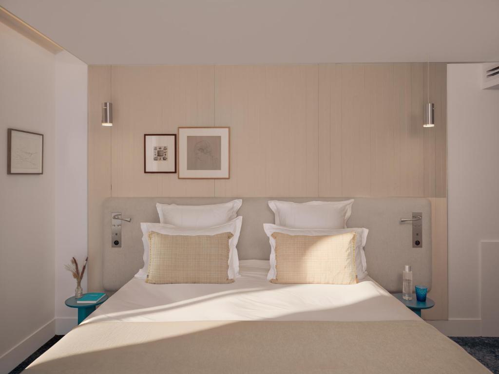 Hotel group accommodation bedroom with a double bed dressed in white linen and two beige woven fringed cushions against a light wood-panelled headboard wall, two chrome reading sconces and two small framed artworks above, small teal round bedside tables with a glass vase of dried grasses on the left and a blue glass on the right, and white walls on each side.