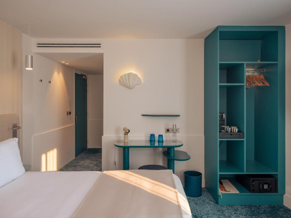 Hotel group accommodation bedroom detail with a teal open wardrobe unit with hanging rail, coffee machine, and safe on the right, a small teal-legged glass-topped console table with two blue glasses and a small lamp in the centre, a white decorative shell wall sconce above, a teal bin on a blue patterned carpet, and an entrance corridor with a teal door visible in the background on the left.