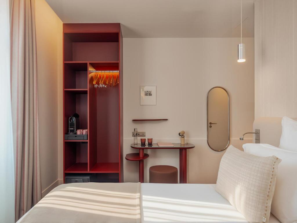 Hotel group accommodation bedroom detail with a red open wardrobe unit with lit hanging rail and a coffee machine on a lower shelf on the left, a small red desk with a round pouf stool and two dark red glasses in the centre, an oval gold-framed mirror and a chrome pendant light on the right wall, a bed with white linen and a beige woven cushion in the foreground, and beige curtains on the left.