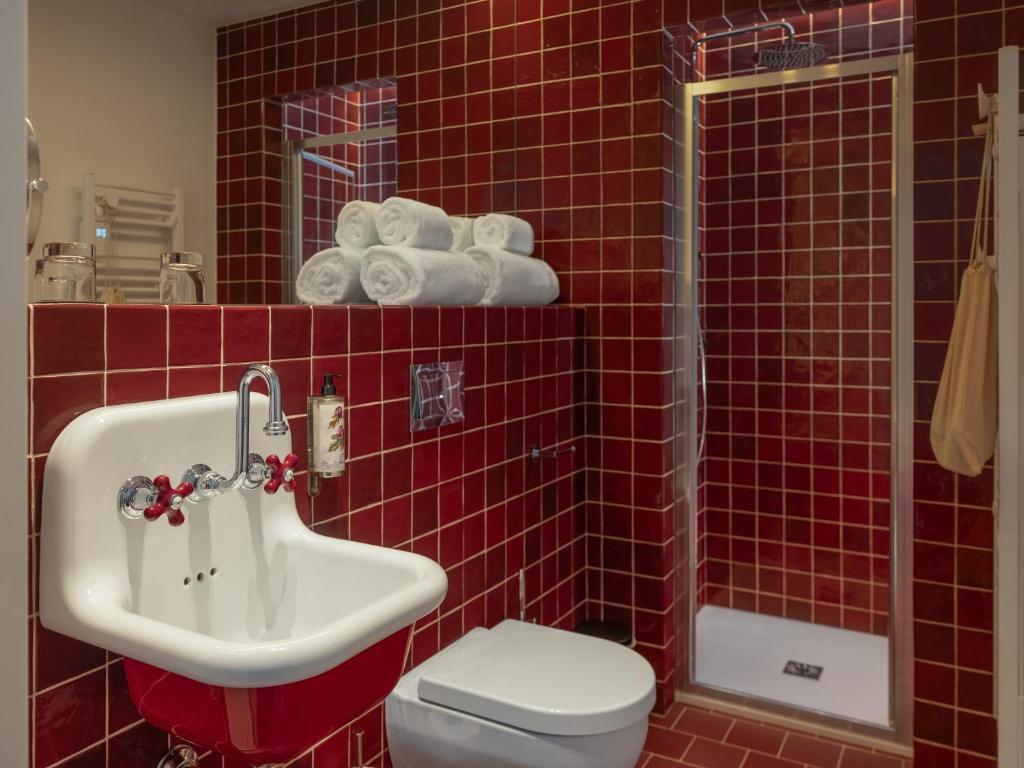 Hotel group accommodation bathroom fully tiled in deep red square tiles, with a vintage white wall-mounted sink with red ceramic tap handles and chrome fittings on the left, rolled white towels stacked on a shelf above a wall mirror in the centre, a wall-hung white toilet, and a glass-door shower cubicle with a chrome overhead fitting on the right.