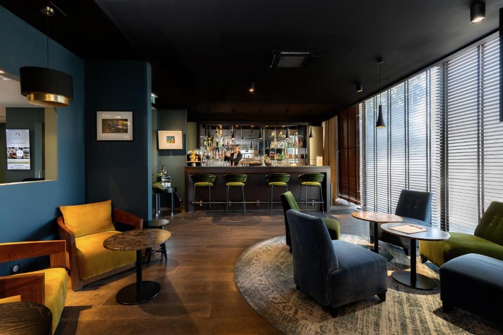 Hotel afterwork bar lounge with a black ceiling and dark parquet floor, mustard yellow armchairs and small round marble-top tables on the left against a teal blue wall, a navy blue and green velvet seating area on a round patterned rug in the centre, a dark wood bar counter with green velvet stools and a bartender in the background, and wood horizontal blinds filtering light from tall windows on the right.