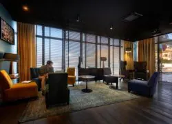 Hotel lobby with a dark ceiling and dark wood floor, featuring an assortment of armchairs in orange, yellow, green, grey and navy arranged around small round tables on a vintage patterned rug, large windows with wooden blinds and ochre curtains letting in natural light, a framed artwork on the blue left wall, and a person seated and reading in the background.