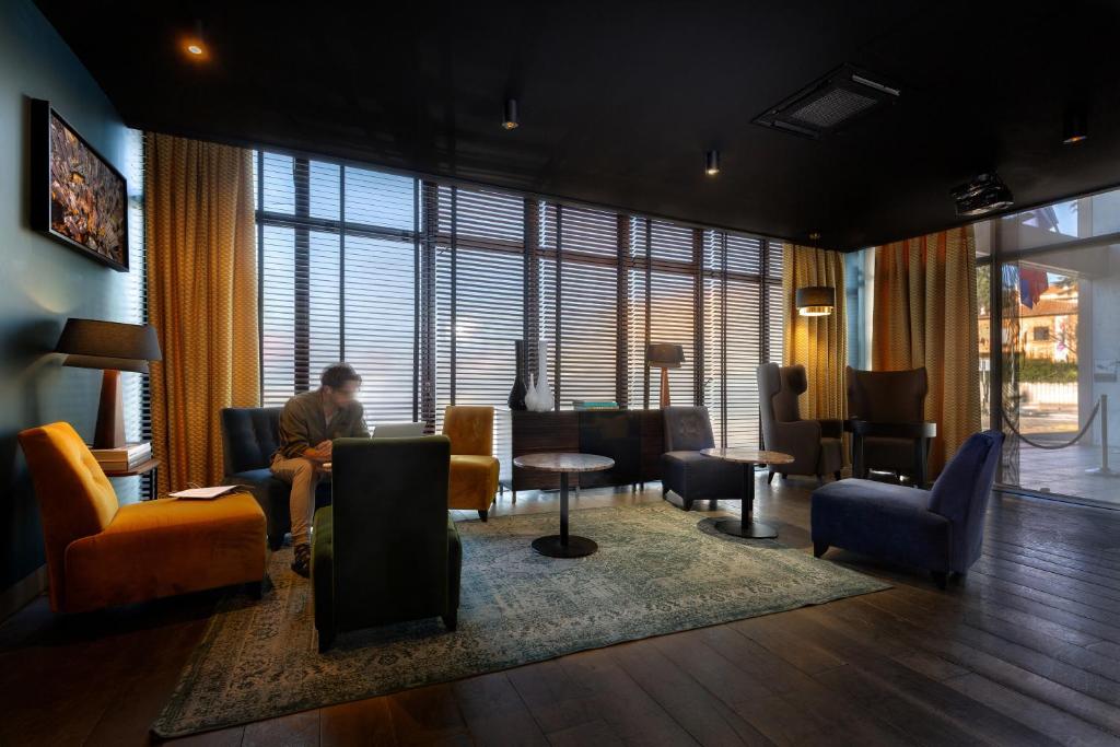 Hotel lobby with a dark ceiling and dark wood floor, featuring an assortment of armchairs in orange, yellow, green, grey and navy arranged around small round tables on a vintage patterned rug, large windows with wooden blinds and ochre curtains letting in natural light, a framed artwork on the blue left wall, and a person seated and reading in the background.
