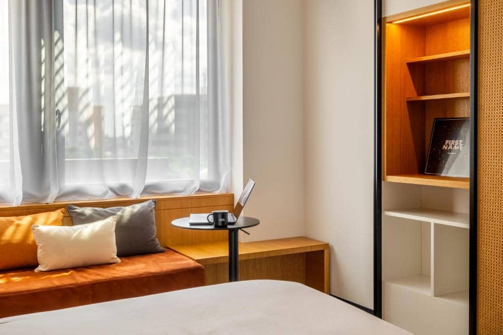 **en::** Bright hotel room with a bed in the foreground, a wooden window bench topped with rust-colored and grey cushions beneath sheer white curtains, and a black round table holding an open laptop; on the right, an illuminated wooden shelving unit adds a warm touch. | **fr::** Chambre d’hôtel lumineuse avec un lit au premier plan, une banquette en bois garnie de coussins brun orangé et gris sous des rideaux blancs translucides, et une table ronde noire accueillant un ordinateur portable ouvert ; à droite, une étagère en bois éclairée apporte une touche chaleureuse.