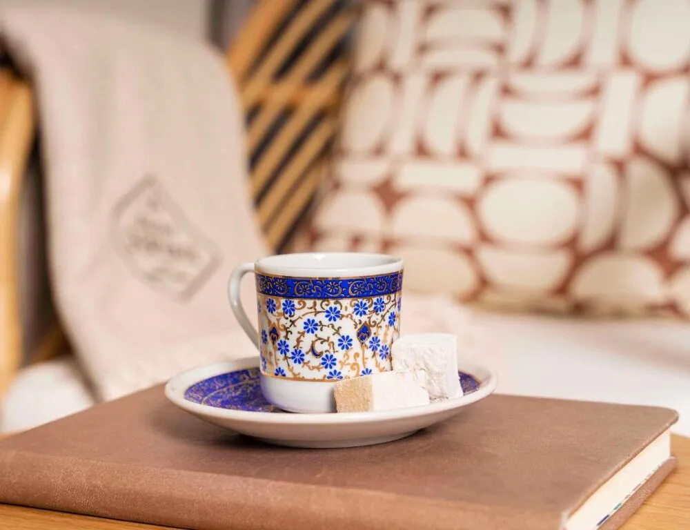 Close-up of an ornate white, blue and gold coffee cup placed on a matching saucer atop a brown book, with pieces of Turkish delight in and beside the cup in the foreground; the softly blurred background reveals a patterned cushion and a light-colored throw draped over an armchair, creating a warm and cozy atmosphere.