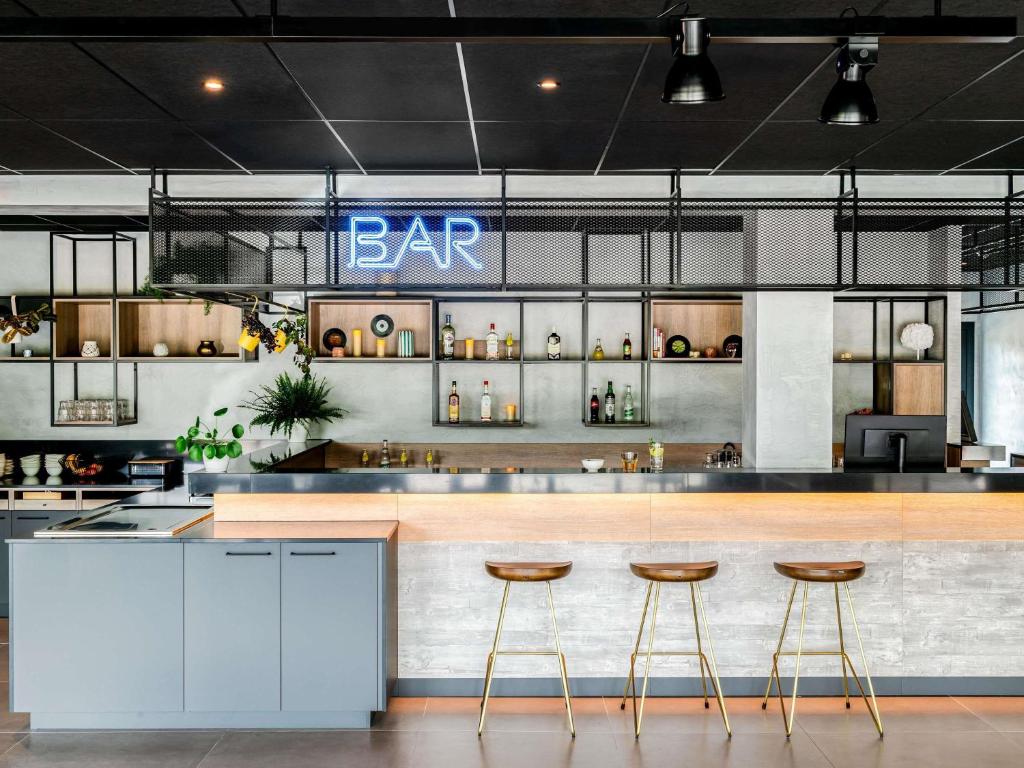 Hotel bar with a black metal mesh ceiling and industrial pendant lights, featuring a blue neon "BAR" sign above open black metal and wood shelving units with bottles and decorative objects, a grey and wood backlit counter with three wooden bar stools on gold legs in the foreground, and grey cabinetry on the left, for an afterwork drink.