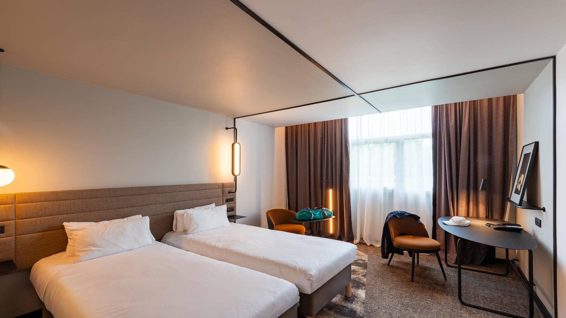 Chambre Novotel spacieuse et très design avec lits twin, tête de lit en tissu marron avec prises et luminaires intégrés, et moquette marron au sol, idéale pour un séjour de groupes à Saint-Quentin-en-Yvelines.