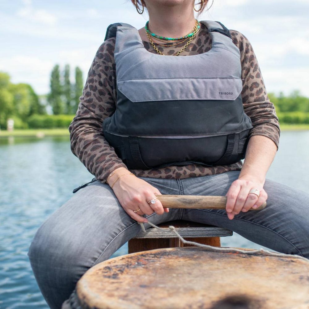 Gros plan d'une personne participant à un séminaire bien-être nautique, portant un gilet de sauvetage gris et noir par-dessus un haut imprimé léopard et un jean gris, tenant un manche de pagaie en bois au premier plan assise sur un bateau, avec une rivière calme et des berges bordées d'arbres verts sous un ciel partiellement nuageux en arrière-plan.