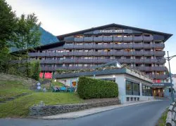 Façade extérieure d'un hôtel pour séminaire à la montagne au crépuscule avec un grand bâtiment chalet multi-étages en bois sombre et gris aux balcons éclairés à encadrements rouges et jardinières fleuries, une entrée illuminée "Chalet Hôtel Le Prieuré" à droite, une petite terrasse extérieure avec chaises bleu canard et clients sur un talus herbeux à gauche, des marches en bois sur le flanc de colline, et une montagne boisée visible sous un ciel bleu pâle de soirée.