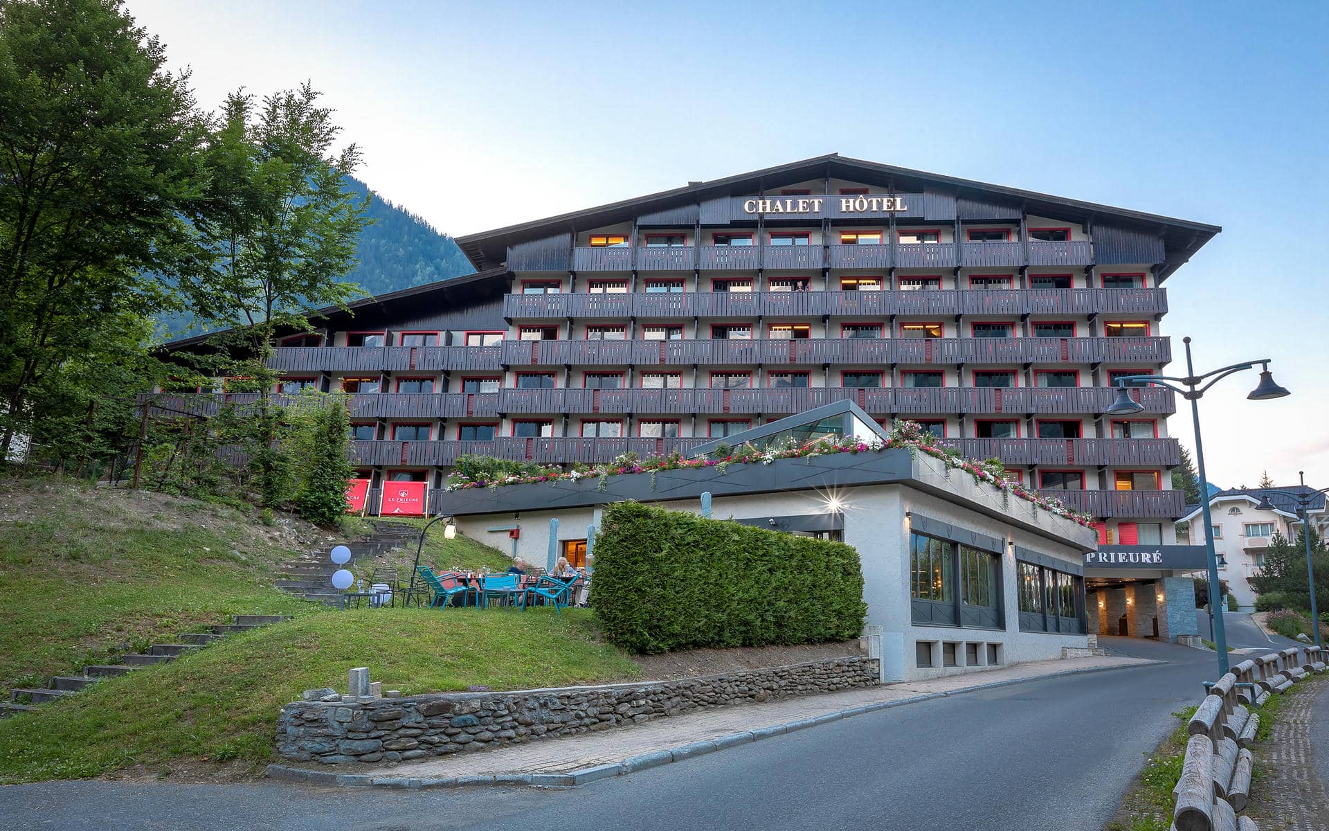 Façade extérieure d'un hôtel pour séminaire à la montagne au crépuscule avec un grand bâtiment chalet multi-étages en bois sombre et gris aux balcons éclairés à encadrements rouges et jardinières fleuries, une entrée illuminée "Chalet Hôtel Le Prieuré" à droite, une petite terrasse extérieure avec chaises bleu canard et clients sur un talus herbeux à gauche, des marches en bois sur le flanc de colline, et une montagne boisée visible sous un ciel bleu pâle de soirée.