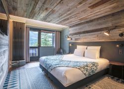 Mountain hotel group accommodation bedroom with weathered grey wood plank walls and ceiling, featuring a double bed with white linens and a teal patterned throw in the centre right, industrial-style swing-arm wall lamps, a dark grey upholstered headboard panel, a wall-mounted TV on the left, a tall black vertical radiator beside a glass door opening onto a balcony with mountain and pine tree views, and a geometric blue and white area rug on the floor.