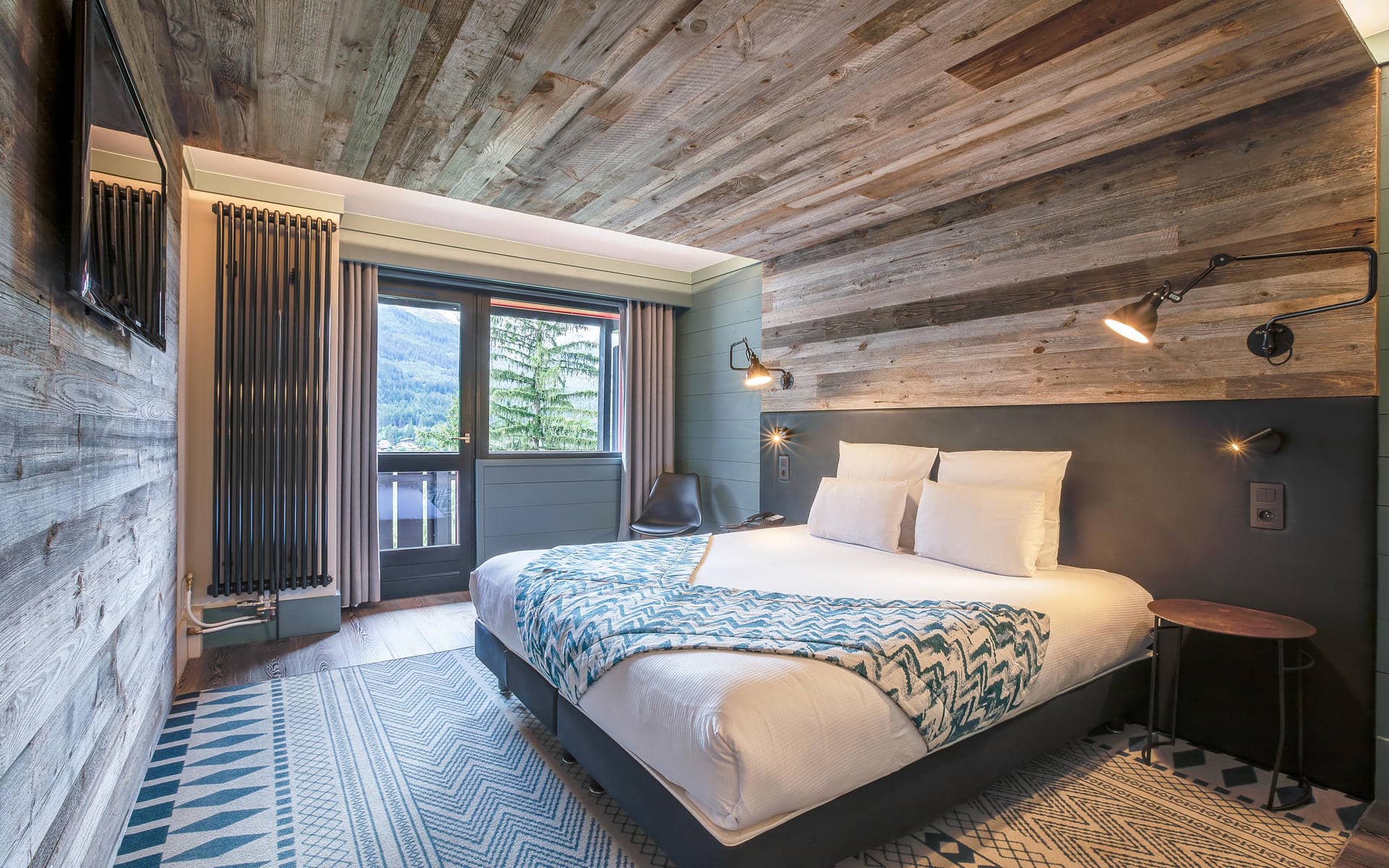 Chambre d'hébergement groupe d'hôtel de montagne avec murs et plafond en planches de bois gris patiné, comprenant un lit double avec linge blanc et jeté à motifs bleu canard au centre droit, des appliques murales articulées de style industriel, une tête de lit en panneau gris foncé, une télévision murale à gauche, un grand radiateur vertical noir près d'une porte vitrée ouvrant sur un balcon avec vue sur montagnes et sapins, et un tapis à motifs géométriques bleu et blanc au sol.