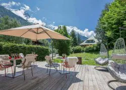 Terrasse extérieure d'hôtel pour séminaire à la montagne avec un deck en bois clair, des chaises en métal rouge drapées de peaux de mouton blanches autour de tables blanches dressées de verres à vin sous un grand parasol beige à gauche, deux fauteuils suspendus en rotin gris avec coussins à droite, une pelouse verte et des haies en arrière-plan, et une vue panoramique sur des sommets enneigés et des versants boisés sous un ciel bleu avec nuages.