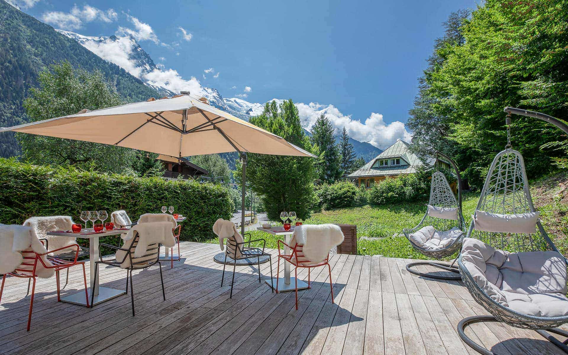 Terrasse extérieure d'hôtel pour séminaire à la montagne avec un deck en bois clair, des chaises en métal rouge drapées de peaux de mouton blanches autour de tables blanches dressées de verres à vin sous un grand parasol beige à gauche, deux fauteuils suspendus en rotin gris avec coussins à droite, une pelouse verte et des haies en arrière-plan, et une vue panoramique sur des sommets enneigés et des versants boisés sous un ciel bleu avec nuages.