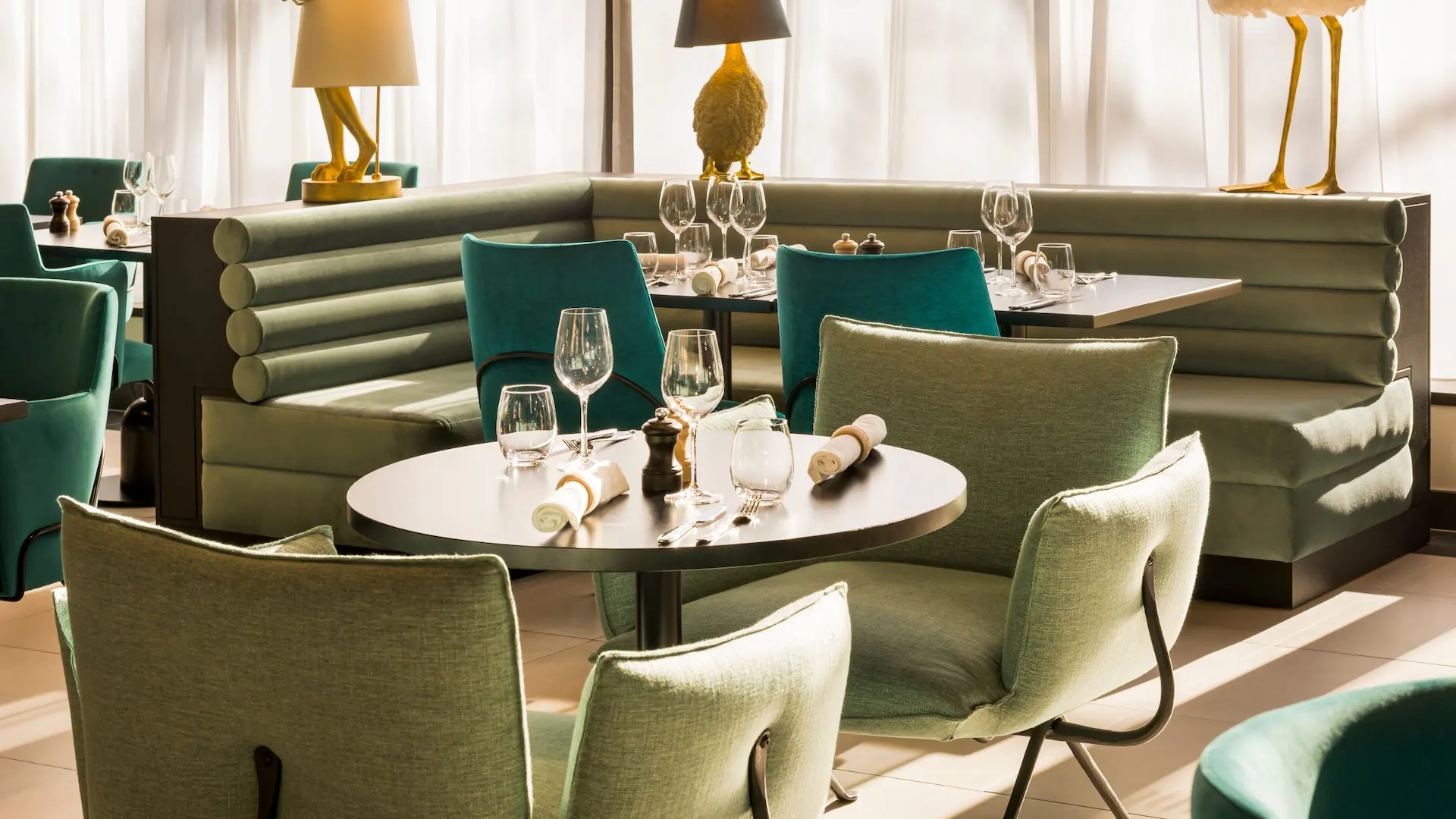 Restaurant dining area in a medium-wide view, featuring in the foreground a round dark table surrounded by light green upholstered armchairs, while in the center and background green banquette seating and turquoise chairs frame neatly set tables with glassware; large sheer-curtained windows and decorative animals-shaped lamps create a bright, elegant atmosphere.
