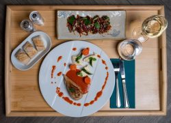 Overhead view of a wooden room service tray on a dark herringbone floor, featuring a white plate with a meat dish in red sauce and vegetable garnish, a rectangular ceramic plate with a starter topped with fresh herbs in the background, a small plate of sliced bread on the left, a glass of white wine and a water glass with a teal napkin and silver cutlery on the right, and salt and pepper shakers in the corner.