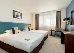 Hotel twin room with grey carpet and blue accent walls, featuring two single beds with white bedding and navy and yellow patterned cushions against a teal headboard panel, a small table with a chair between the beds, a window with taupe curtains in the centre, and a wooden desk with a lamp and wall-mounted television on the right, for group accommodation.