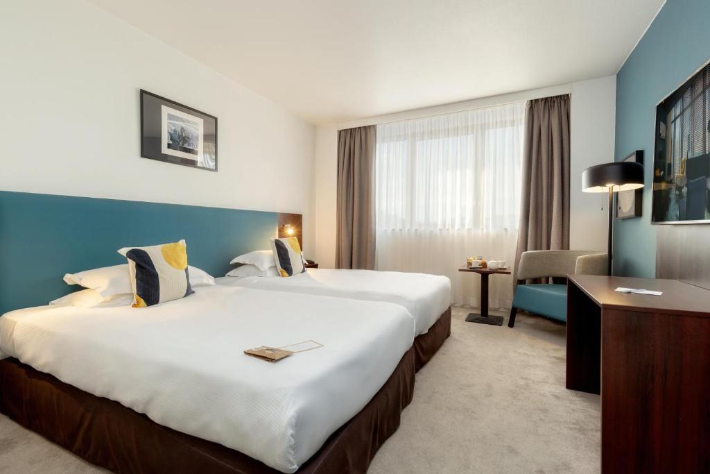 Hotel twin room with grey carpet and blue accent walls, featuring two single beds with white bedding and navy and yellow patterned cushions against a teal headboard panel, a small table with a chair between the beds, a window with taupe curtains in the centre, and a wooden desk with a lamp and wall-mounted television on the right, for group accommodation.