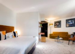 Spacious hotel room with light grey carpet and white walls, featuring a double bed with an ochre velvet headboard and navy and orange patterned cushions on the left, an open metal shelving unit with a safe in the centre, and a mustard yellow sofa with a glass coffee table, an orange armchair and two framed artworks on the wall on the right, for group accommodation.