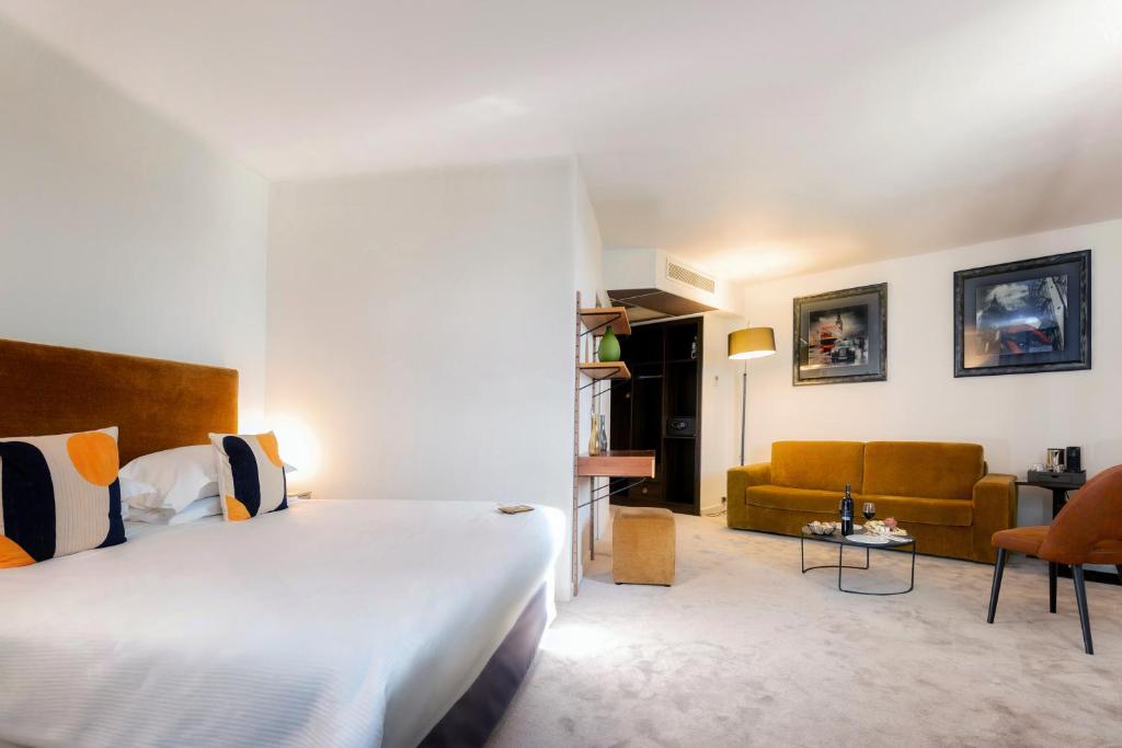 Spacious hotel room with light grey carpet and white walls, featuring a double bed with an ochre velvet headboard and navy and orange patterned cushions on the left, an open metal shelving unit with a safe in the centre, and a mustard yellow sofa with a glass coffee table, an orange armchair and two framed artworks on the wall on the right, for group accommodation.