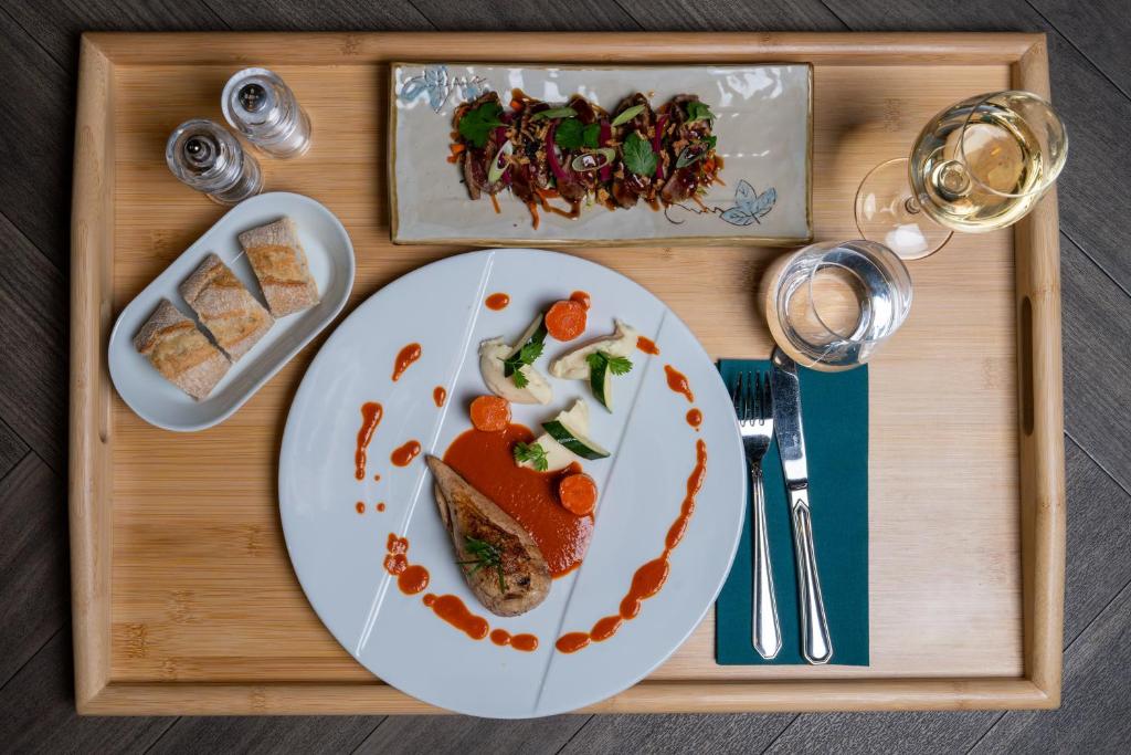 Overhead view of a wooden room service tray on a dark herringbone floor, featuring a white plate with a meat dish in red sauce and vegetable garnish, a rectangular ceramic plate with a starter topped with fresh herbs in the background, a small plate of sliced bread on the left, a glass of white wine and a water glass with a teal napkin and silver cutlery on the right, and salt and pepper shakers in the corner.