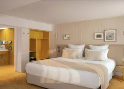 Hotel bedroom with a double bed dressed in white linens occupying the foreground on the right, featuring a bright yellow open wardrobe at the center-left, and an open bathroom area with mustard-yellow tiled walls and a vintage-style wall-mounted sink on the far left; light wood wall paneling, framed artwork, and soft neutral tones create a contemporary and inviting atmosphere.