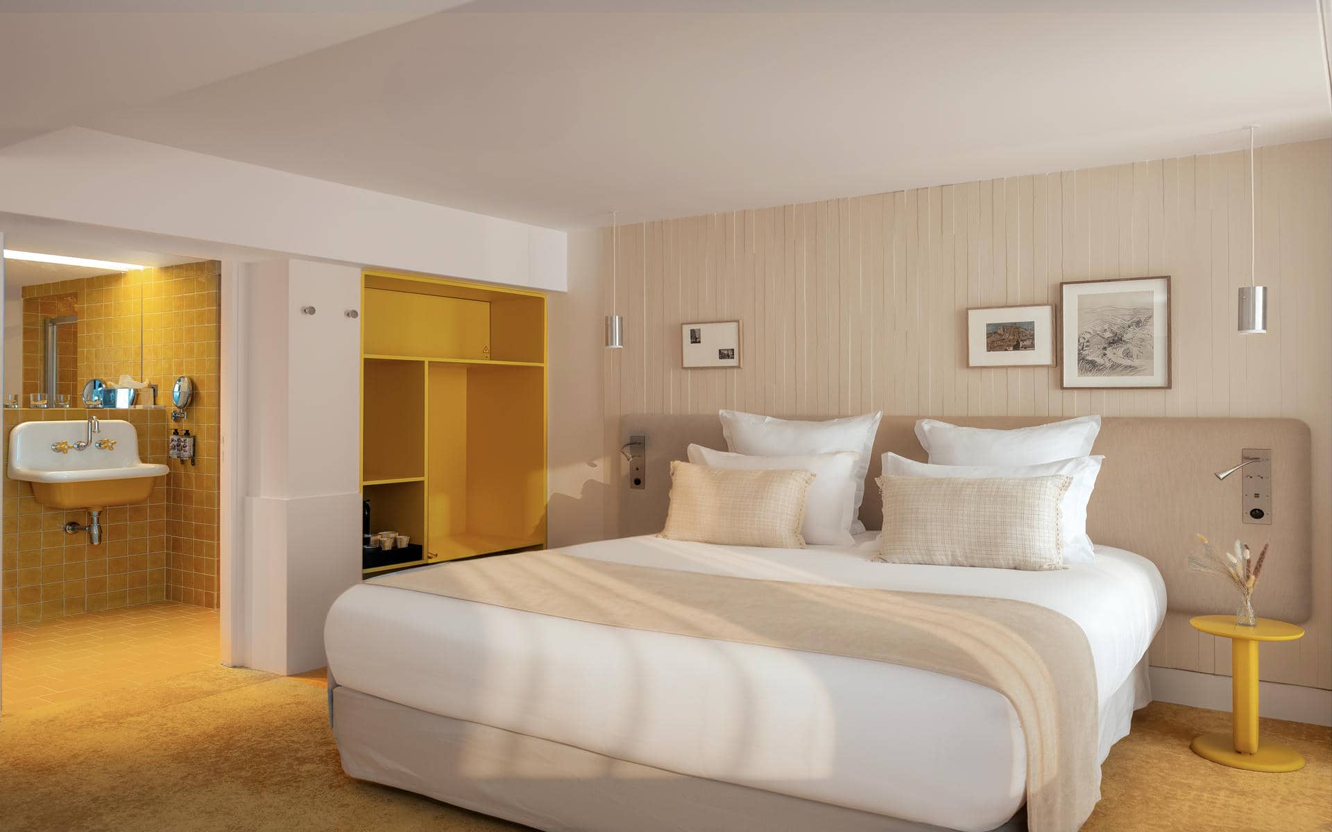Hotel bedroom with a double bed dressed in white linens occupying the foreground on the right, featuring a bright yellow open wardrobe at the center-left, and an open bathroom area with mustard-yellow tiled walls and a vintage-style wall-mounted sink on the far left; light wood wall paneling, framed artwork, and soft neutral tones create a contemporary and inviting atmosphere.