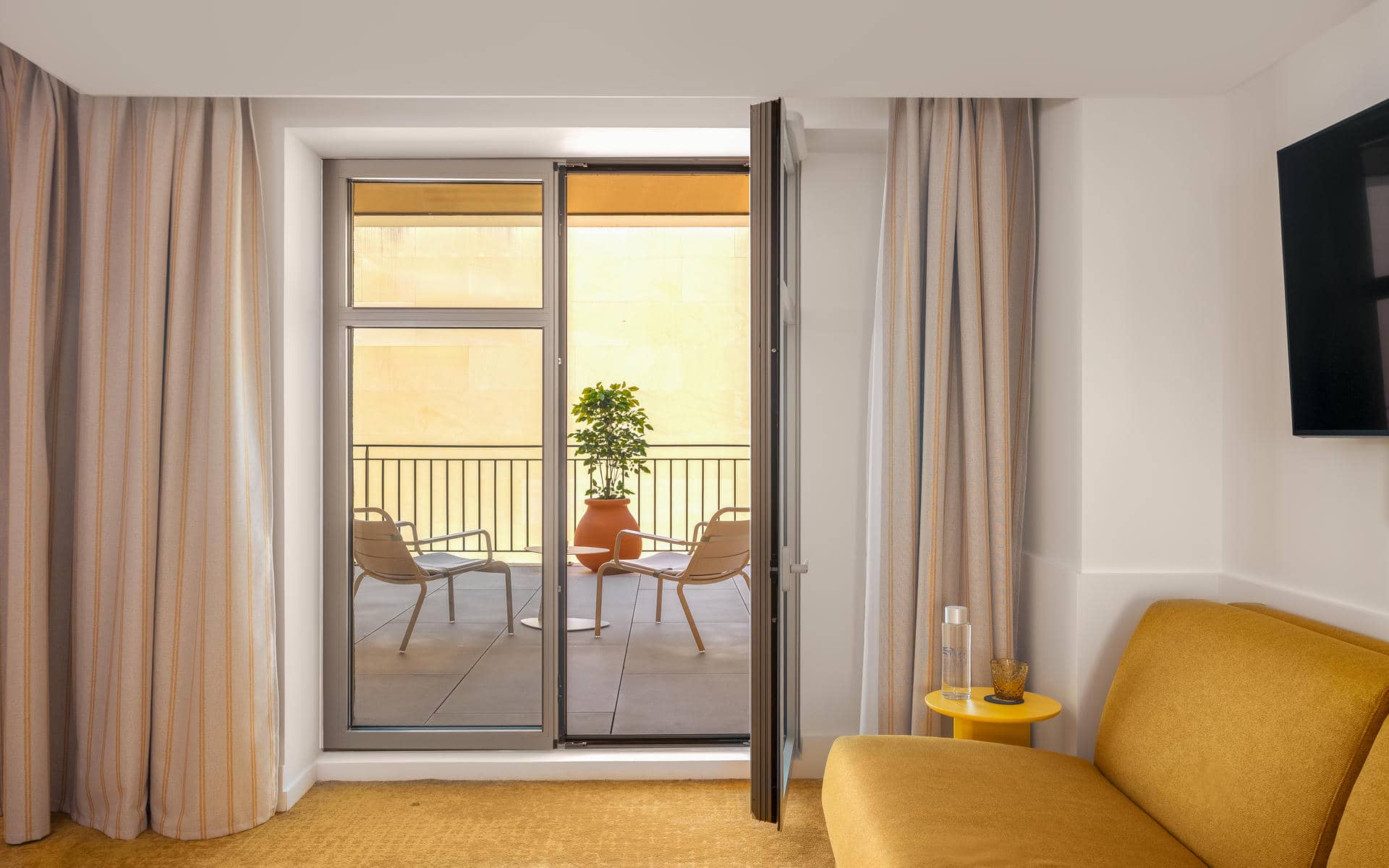 Hotel group accommodation room with a yellow upholstered armchair and a small yellow side table in the foreground on a mustard carpet, beige striped curtains framing an open glass door leading to a balcony with two beige metal chairs, a terracotta pot with a green plant, a grey tiled floor and a metal railing, with a flat screen television on the white wall to the right.