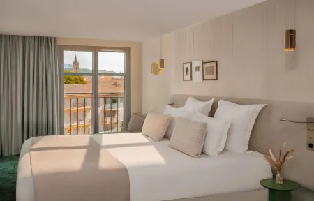 Urban seminar hotel bedroom with a double bed dressed in white linen and beige cushions against a light wood-panelled headboard wall, gold pendant lights and two framed artworks above, a small green round bedside table with dried grasses on the right, a dark green carpet, grey linen curtains framing a window with a view over the Aix-en-Provence rooftops and a church bell tower.