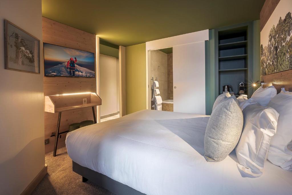 Chambre double d'hôtel avec plafond vert olive et lambris en pin, comprenant un bureau en bois et téléviseur mural avec photographie de montagne à gauche, un passage ouvert vers la salle de bain avec serviettes visibles au centre, et une niche d'étagères bleues avec machine à café et applique de lecture à côté d'une tête de lit murale paysagère à droite, une chambre bien équipée pour un hôtel pour séminaire.