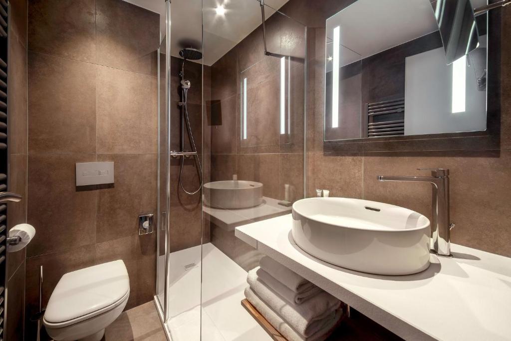 Salle de bain d'hôtel entièrement habillée de grands carreaux effet pierre brun foncé, avec WC suspendu et douche à paroi vitrée à gauche, et un plan vasque blanc avec deux vasques rondes à poser, serviettes pliées en dessous et grand miroir éclairé à droite.