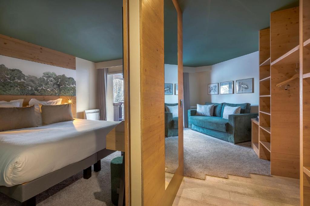 Suite d'hôtel de montagne vue à travers une cloison en pin, avec une chambre comprenant un lit double et une tête de lit murale paysagère à gauche, et un salon avec canapé bleu canard, gravures encadrées et étagères ouvertes à droite, les deux espaces partageant un plafond bleu canard foncé, une disposition confortable pour un séminaire à la montagne.
