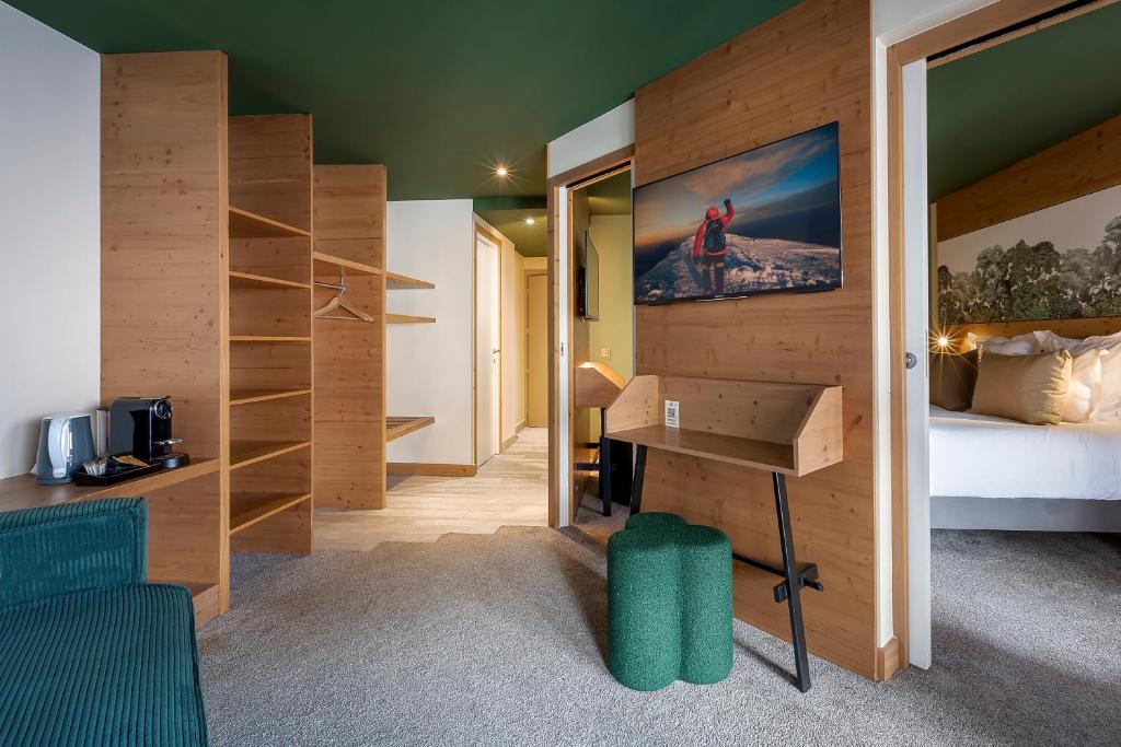 Chambre d'hôtel de montagne avec plafond vert foncé et lambris en pin clair, comprenant des étagères ouvertes et une machine à café à gauche, un téléviseur mural au-dessus d'un bureau en bois avec pouf vert texturé au centre, et un lit avec tête de lit murale paysagère partiellement visible à droite, un espace bien équipé pour un séminaire à la montagne.