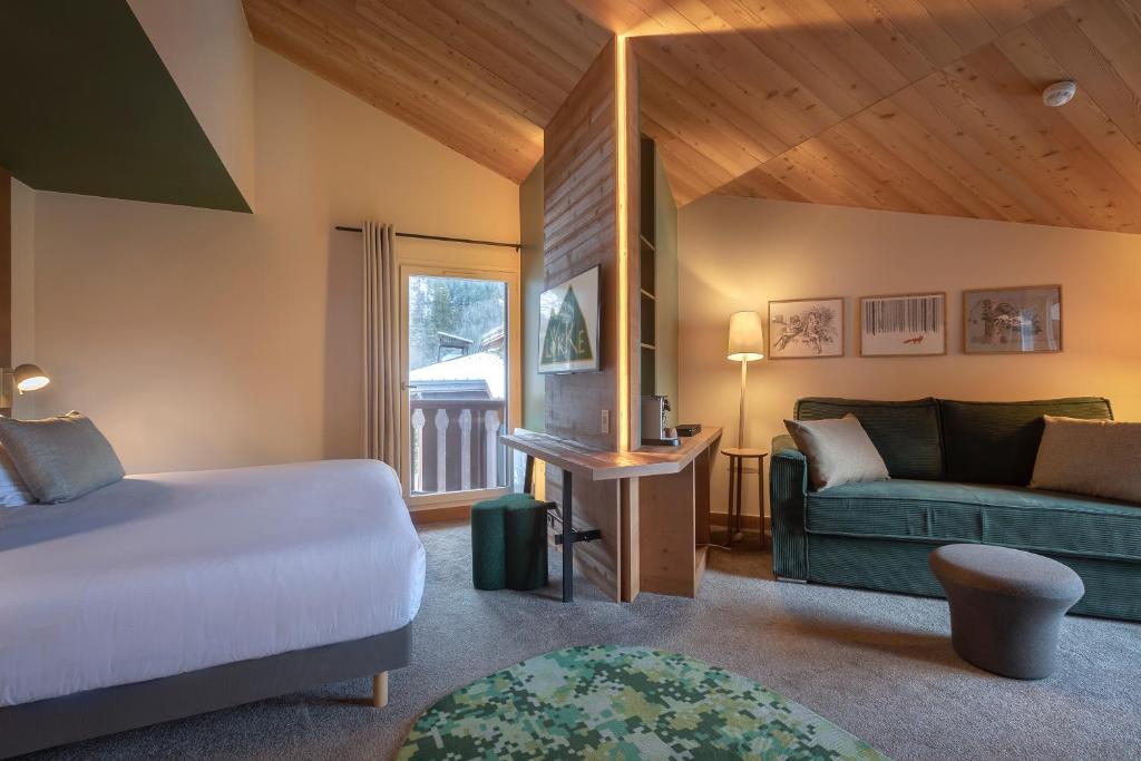 Suite d'hôtel de montagne spacieuse avec plafond en pente en bois de pin, comprenant un lit à gauche, une séparation bureau en bois avec bandeau LED chaud au centre, un canapé en velours vert avec gravures encadrées sur le mur à droite, un tapis rond à motifs au premier plan et une fenêtre donnant sur un balcon enneigé, un espace polyvalent pour un séminaire à la montagne.
