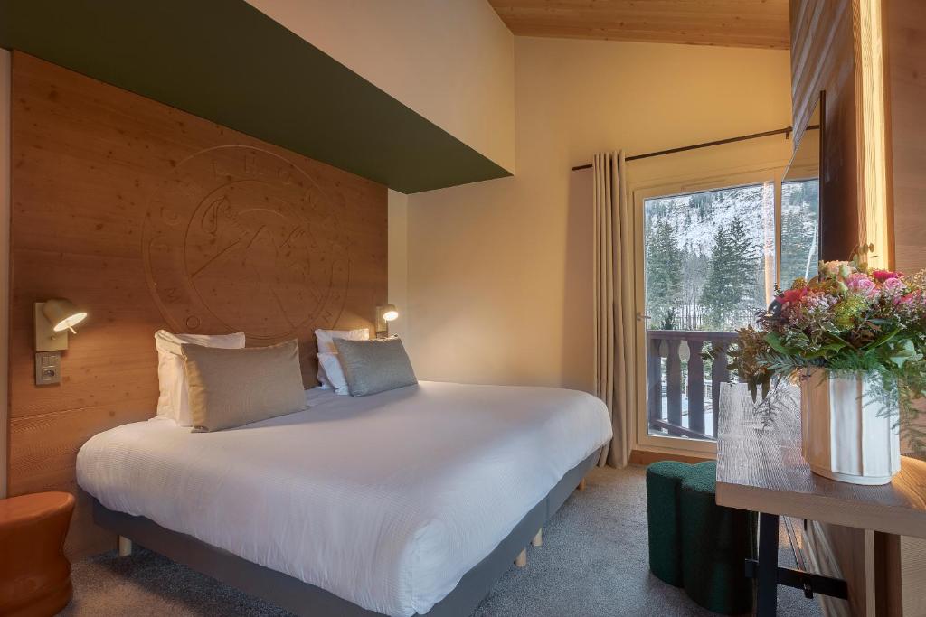 Chambre double d'hôtel de montagne avec une tête de lit en bois sculpté ornée d'un motif alpin gravé, literie grise et appliques de lecture de chaque côté, plafond en pente en bois de pin, et une fenêtre à droite donnant sur une forêt enneigée, avec un bureau en bois supportant un bouquet de fleurs coloré au premier plan, un cadre chaleureux pour un séminaire à la montagne.