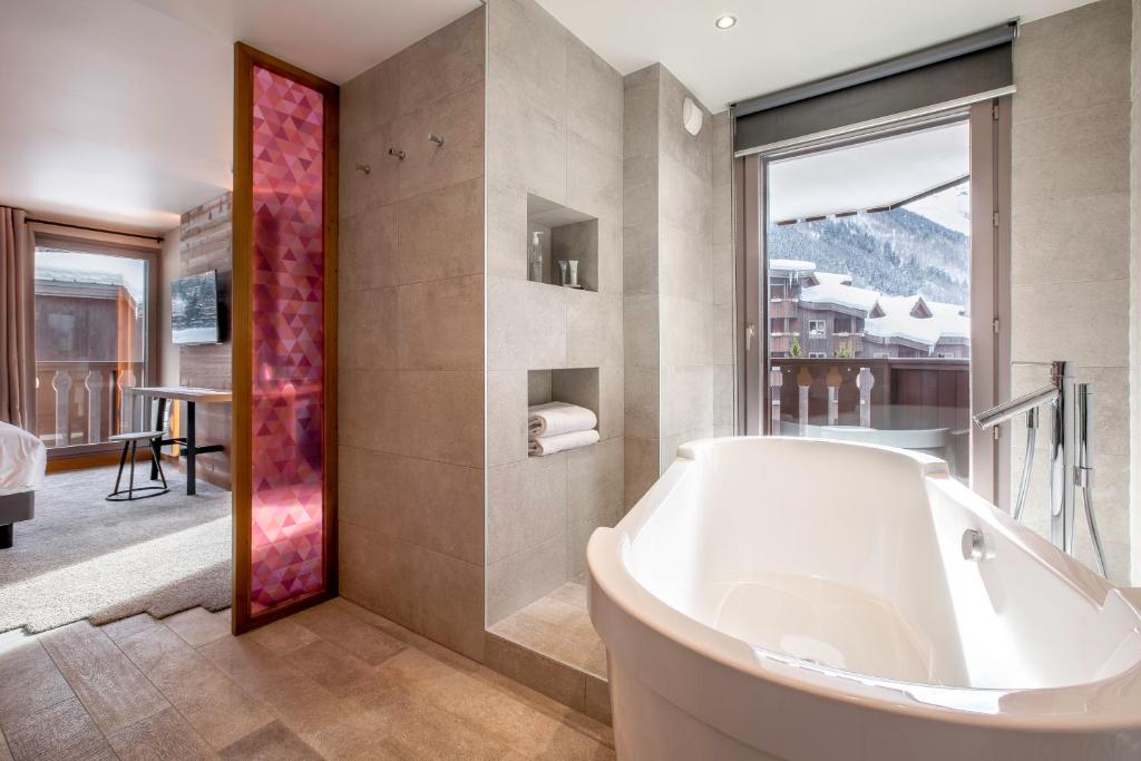 Salle de bain d'hôtel en grands carreaux de pierre grise, avec une baignoire blanche îlot au premier plan à droite face à une fenêtre donnant sur un paysage de montagne enneigé, des niches encastrées avec produits et serviettes au centre, et un remarquable panneau décoratif géométrique rose encadrant le passage vers la chambre à gauche, un cadre raffiné pour un séjour de séminaire à la montagne.