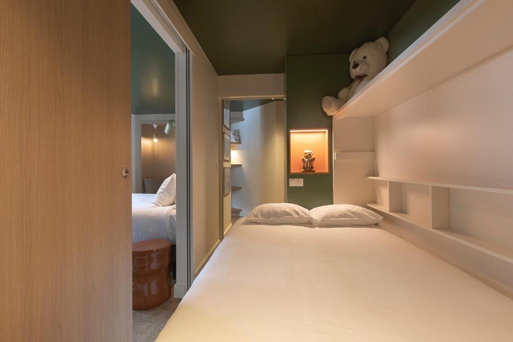 Chambre d'hôtel compacte avec plafond vert foncé et lambris bois clair, comprenant un lit simple en literie blanche et des étagères blanches encastrées avec un ours en peluche et une niche décorative éclairée à droite, et une porte miroir coulissante ouvrant sur une seconde chambre avec lit double et table de chevet vernie en arrière-plan, une configuration pratique pour un hébergement groupe.
