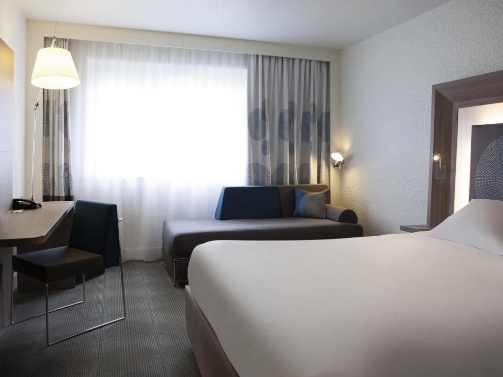 Chambre d'hôtel adaptée pour un hébergement groupe, comprenant un lit blanc au premier plan, un canapé-lit gris et bleu devant un rideau à motifs en arrière-plan, un bureau en bois avec une chaise noire à gauche, et un lampadaire à côté de rideaux blancs voilés sous une lumière naturelle.