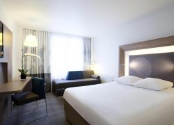 Chambre d'hôtel avec moquette grise à motifs, un bureau en bois avec chaise bleu marine, un lampadaire et une orchidée près de rideaux voilage à gauche, une banquette grise avec coussins bleus et beiges au plan intermédiaire, et un grand lit avec linge blanc adossé à une tête de lit en bois ornée d'un motif circulaire à pois à droite, adaptée à de l'hébergement groupe.
