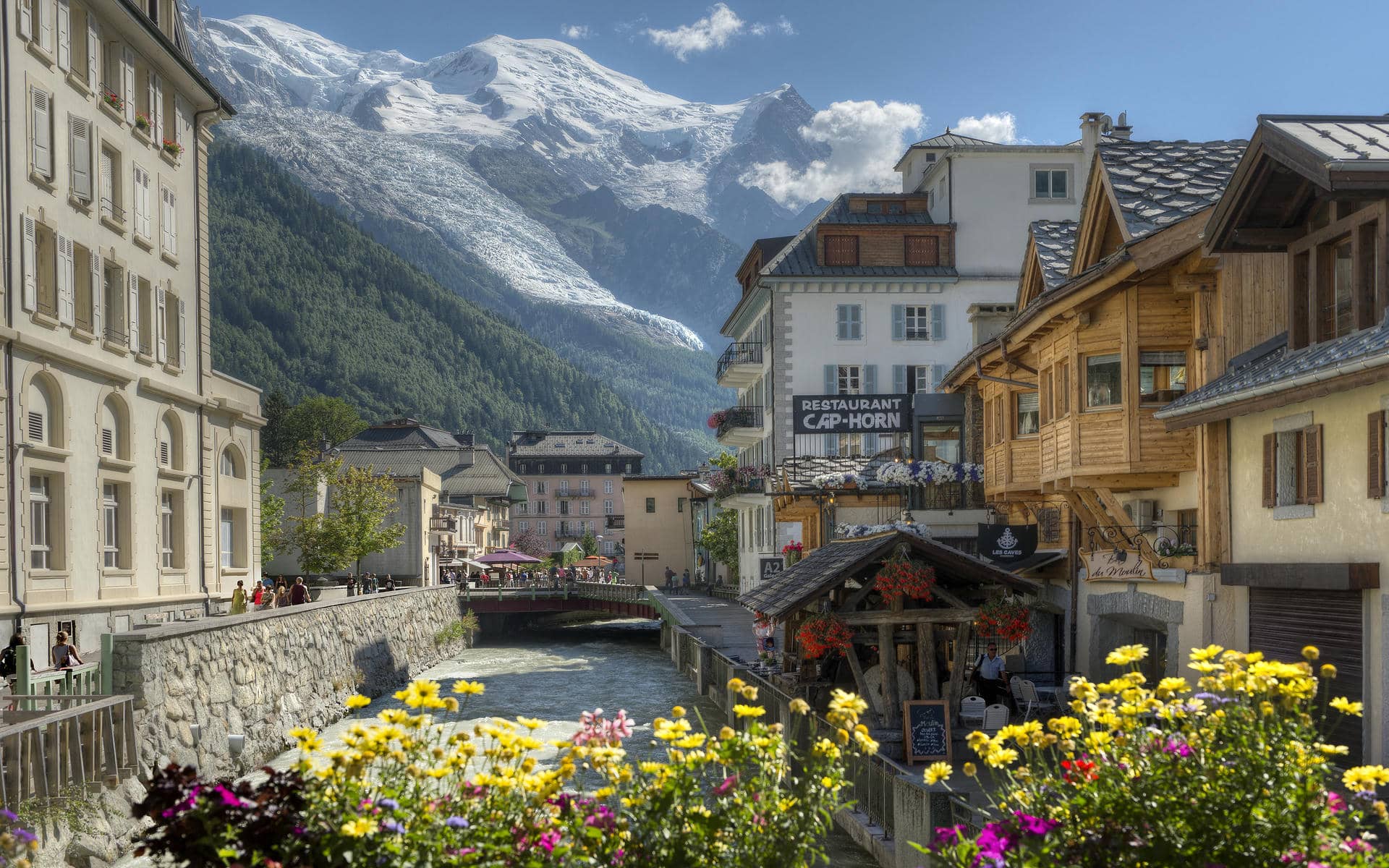 View of Chamonix town center with a mountain river running through the middle, historic buildings on the left and wooden chalet-style façades on the right, colorful flowers in the foreground and snow-capped peaks dominating the background, illustrating a destination for a séminaire à la montagne.
