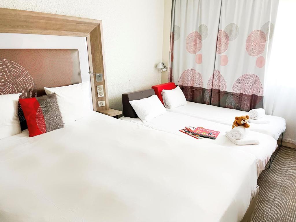 Chambre d'hôtel familiale adaptée pour un hébergement groupe, comprenant un large lit avec tête de lit en bois et coussins rouges à motifs au premier plan, un second lit avec des serviettes blanches roulées, un ours en peluche et un magazine en arrière-plan, et un rideau à motifs roses et gris devant un mur blanc à droite.