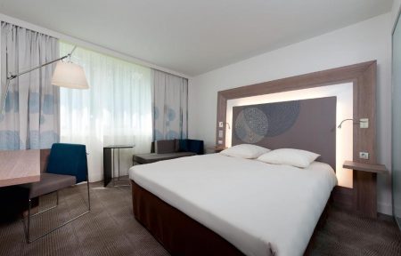 Chambre d'hôtel avec moquette grise à motifs, un bureau avec chaise bleu canard et lampadaire près de rideaux voilage à gauche, et un grand lit avec linge blanc adossé à une tête de lit en bois ornée d'un panneau circulaire à motifs gris et de liseuses intégrées au centre, adaptée à de l'hébergement groupe.