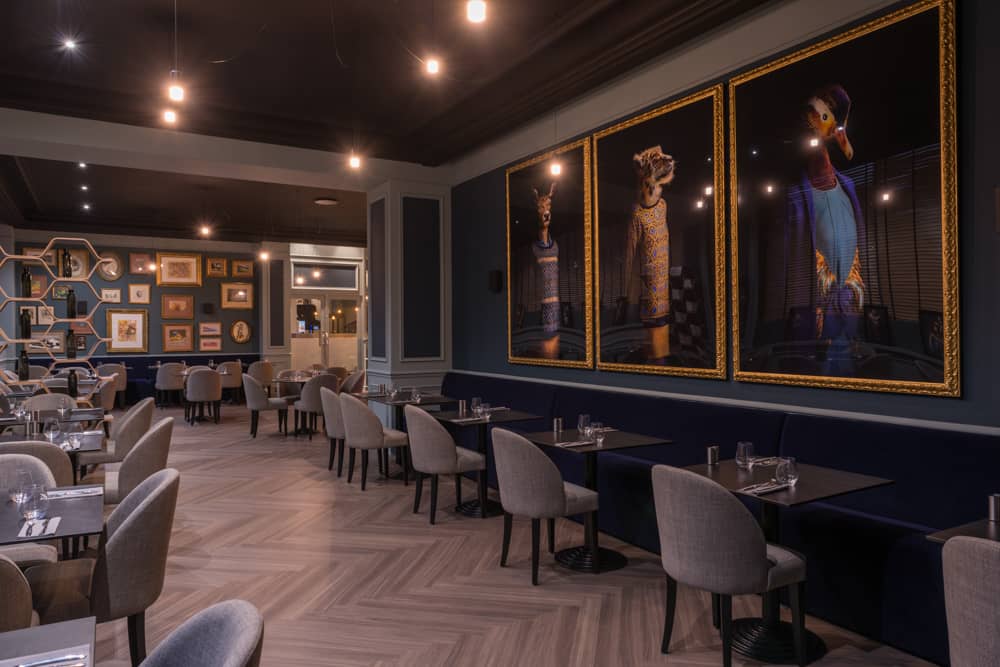 Salle de restauration sur place d'hôtel avec un parquet clair en chevrons, des tables noires et des chaises tapissées grises, trois grandes œuvres contemporaines à cadre doré représentant des animaux habillés sur le mur droit bleu marine foncé, une étagère hexagonale avec de petits cadres sur le mur du fond bleu foncé, et un éclairage chaleureux par suspensions sous un plafond sombre.