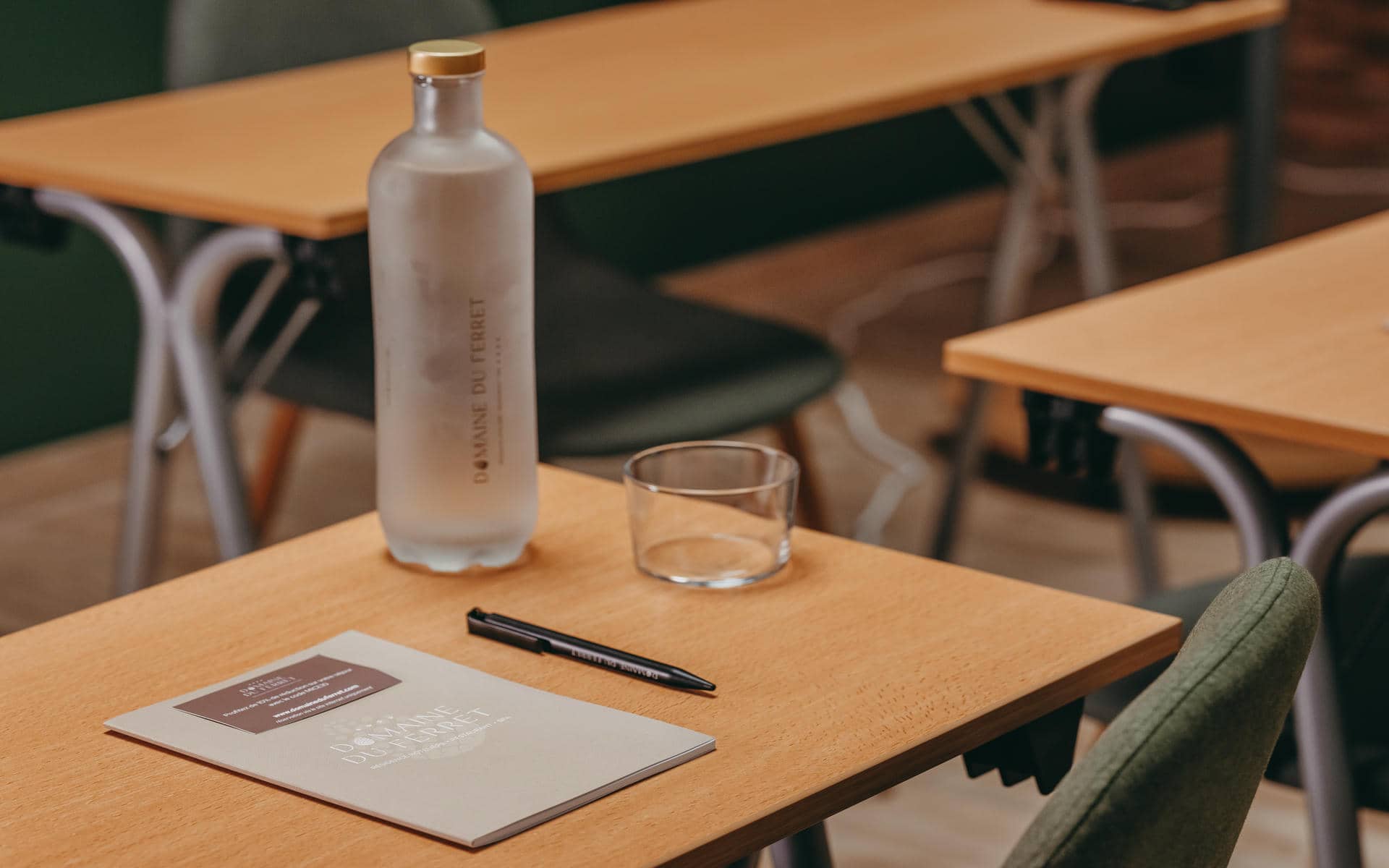 Close-up of a classroom-style meeting table, featuring a frosted glass water bottle, glass tumbler, notebook, and pen arranged in the foreground, with additional tables and chairs softly blurred in the background; light wood surfaces and neutral tones create a professional meeting setting.