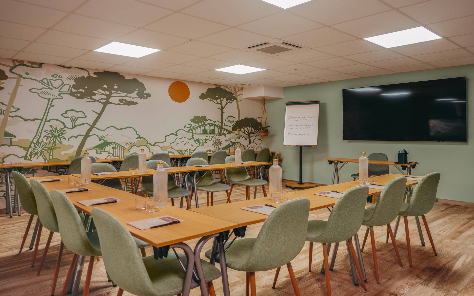 Meeting room arranged in a U-shape, featuring light wood tables surrounded by pale green chairs with notepads and water bottles at each place; in the background, a large wall mural depicting a forest landscape faces a widescreen display and a flip chart, while a wood-look floor completes the space.