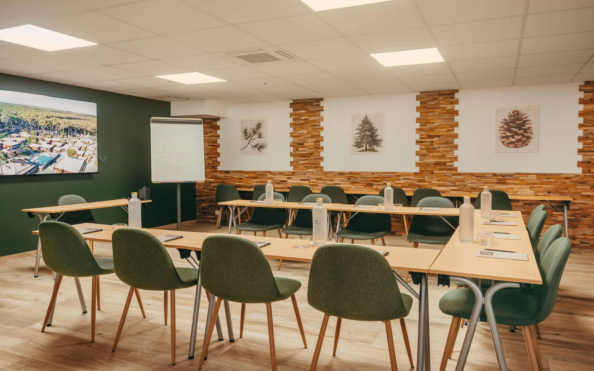Meeting room arranged in a U-shape configuration with light wood flooring, featuring tables equipped with notebooks, glass bottles, and tumblers surrounding the center space, green upholstered chairs throughout, a presentation screen and flip chart on the left, and decorative wood wall panels with nature-themed artwork in the background.