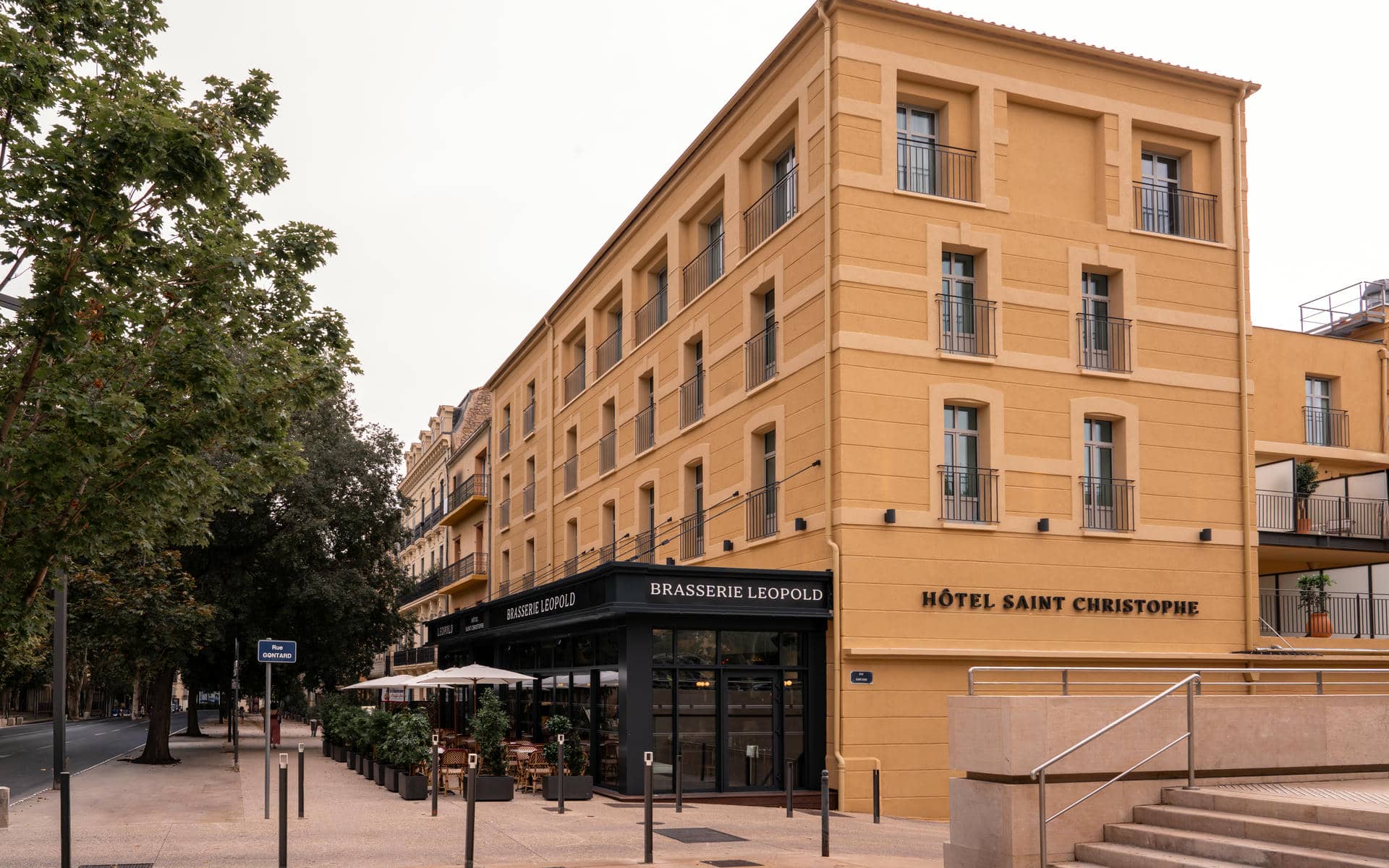Urban seminar hotel exterior with a large ochre-yellow multi-storey Haussmann-style facade with black iron balconies, a black-canopied "Brasserie Léopold" street terrace with white parasols and bistro chairs on the left, the "Hôtel Saint Christophe" signage on the corner, stone steps leading to the entrance on the right, and a tree-lined boulevard under a pale grey sky.